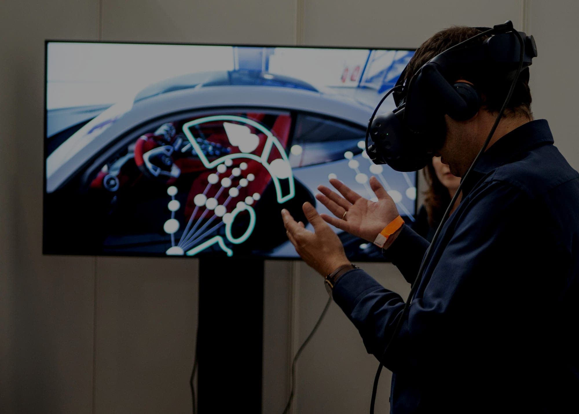A man wearing a VR headset gestures with both hands at a screen showing a car interior overlaid with a glowing interface