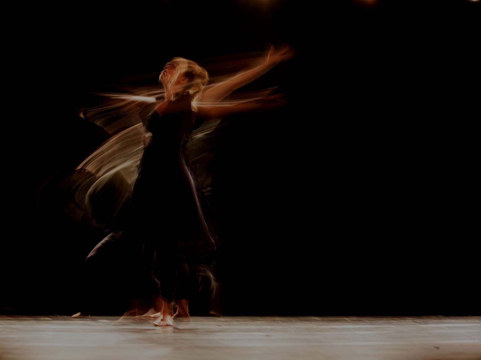 A dancer in a dark dress photographed with long exposure, her movement trailing across a lit stage
