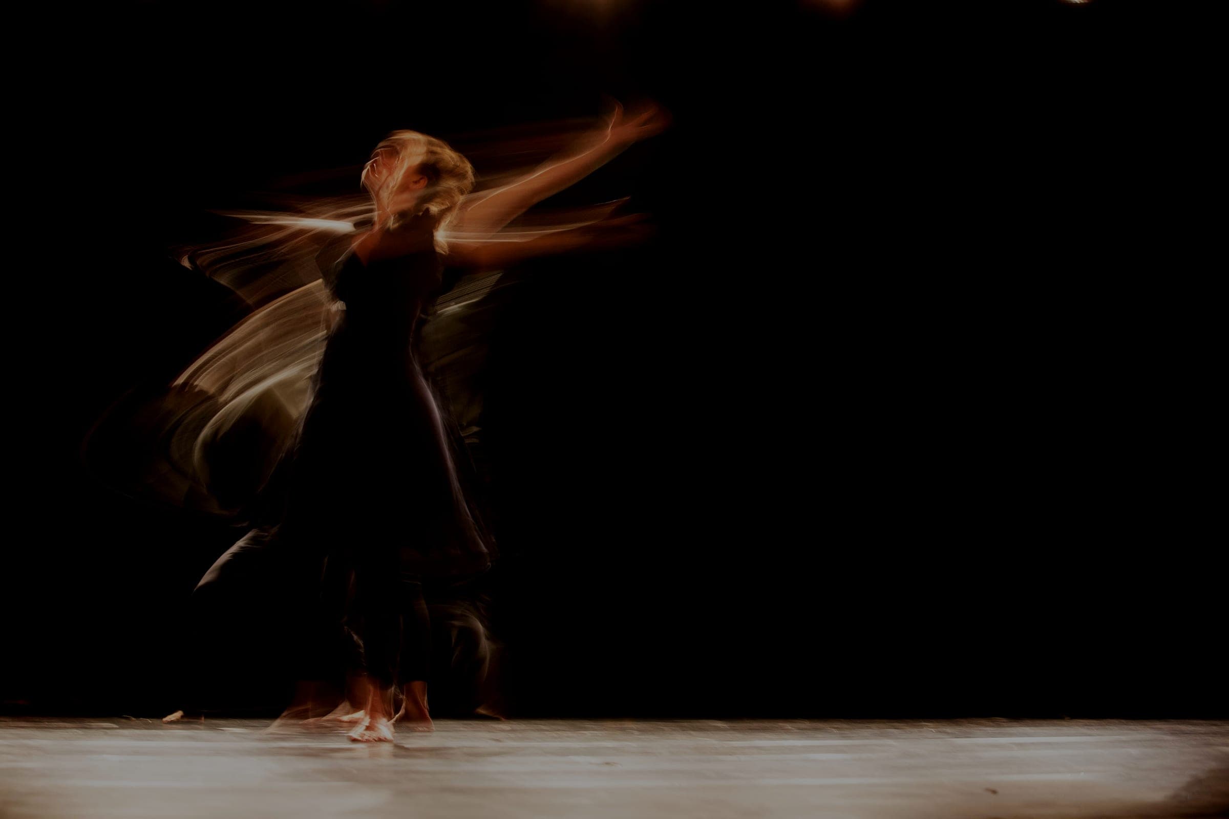 A dancer in a dark dress photographed with long exposure, her movement trailing across a lit stage