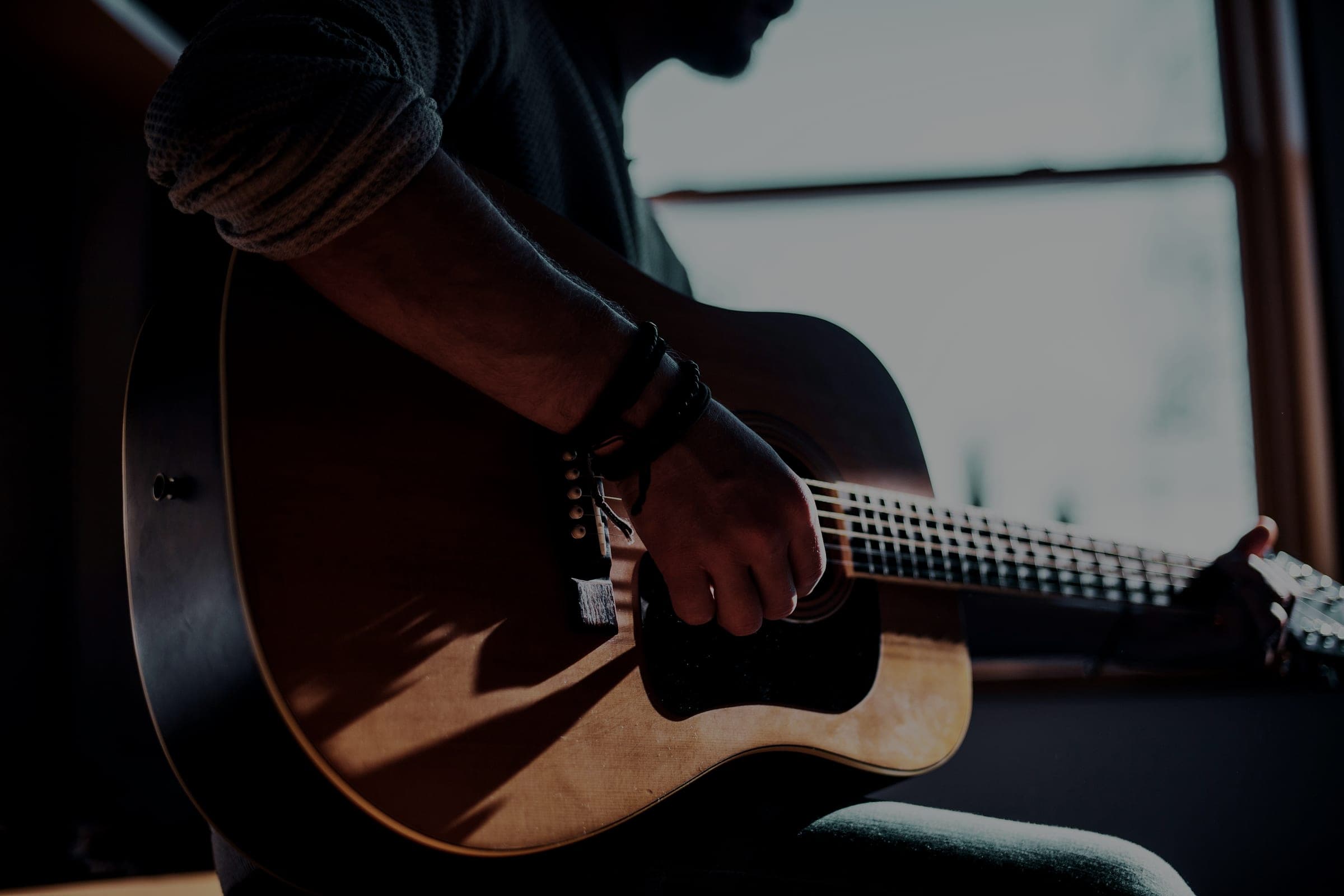 A person playing an acoustic guitar by a window in low winter light