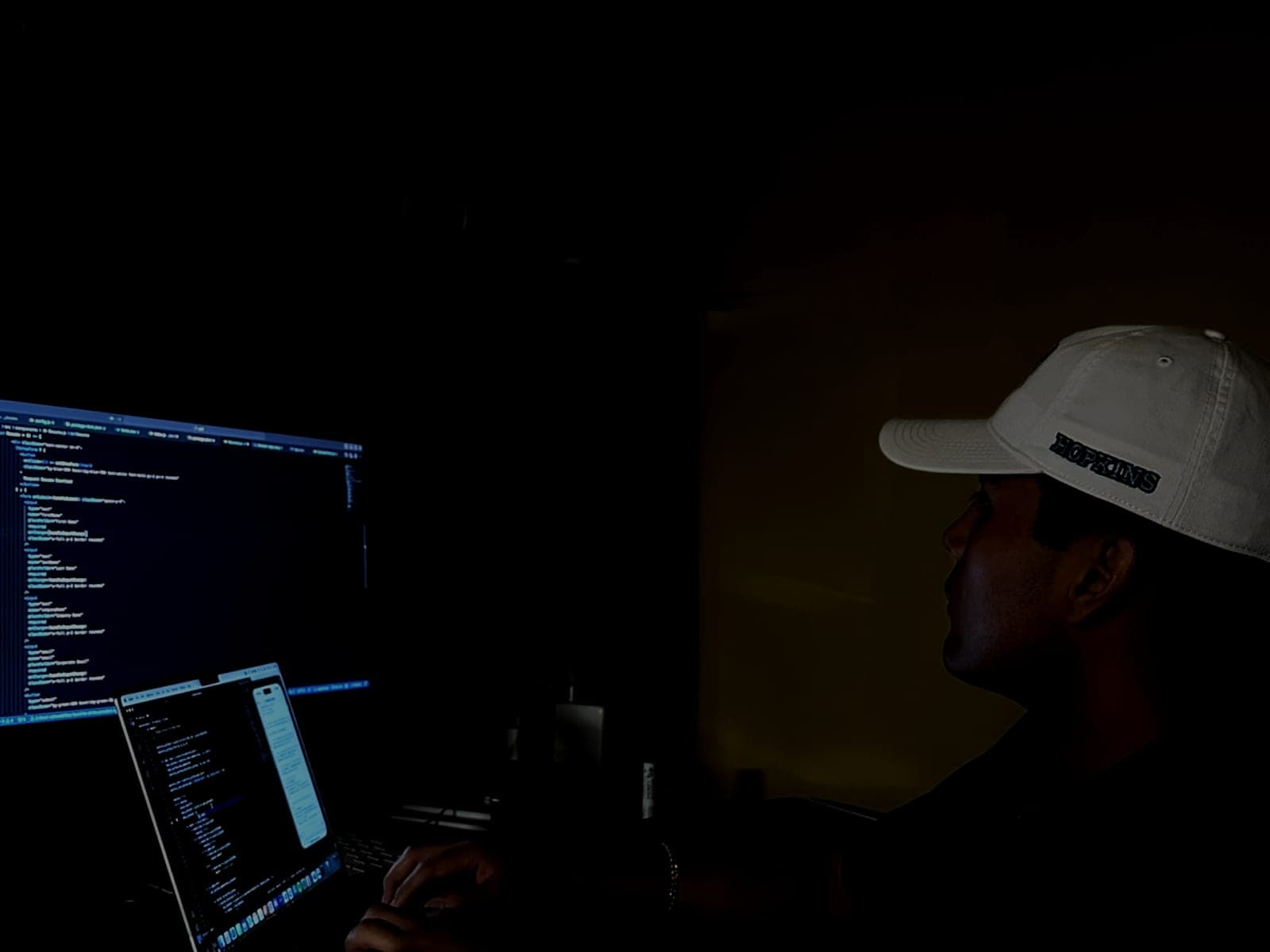 Jeffrey Escobar coding AltaRealm, an AI world-building game, late at night in August 2025, wearing a Johns Hopkins cap