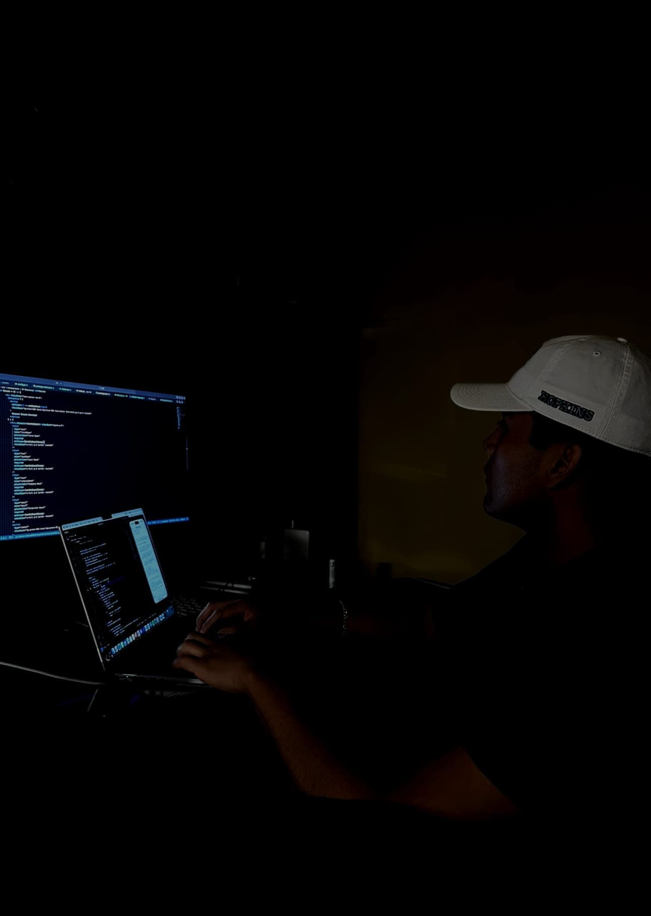 Jeffrey Escobar coding AltaRealm, an AI world-building game, late at night in August 2025, wearing a Johns Hopkins cap
