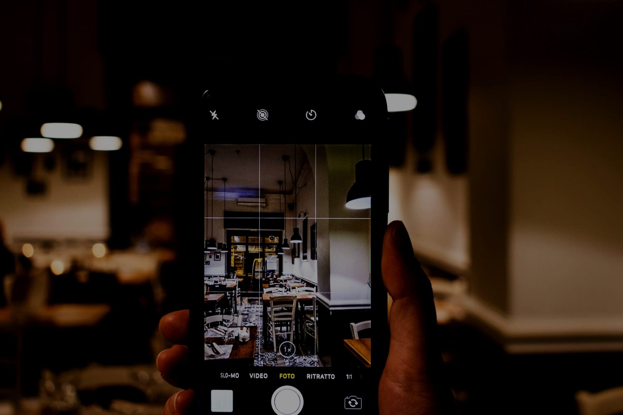 A hand holding a phone in camera mode, framing the tables and pendant lights of a restaurant interior