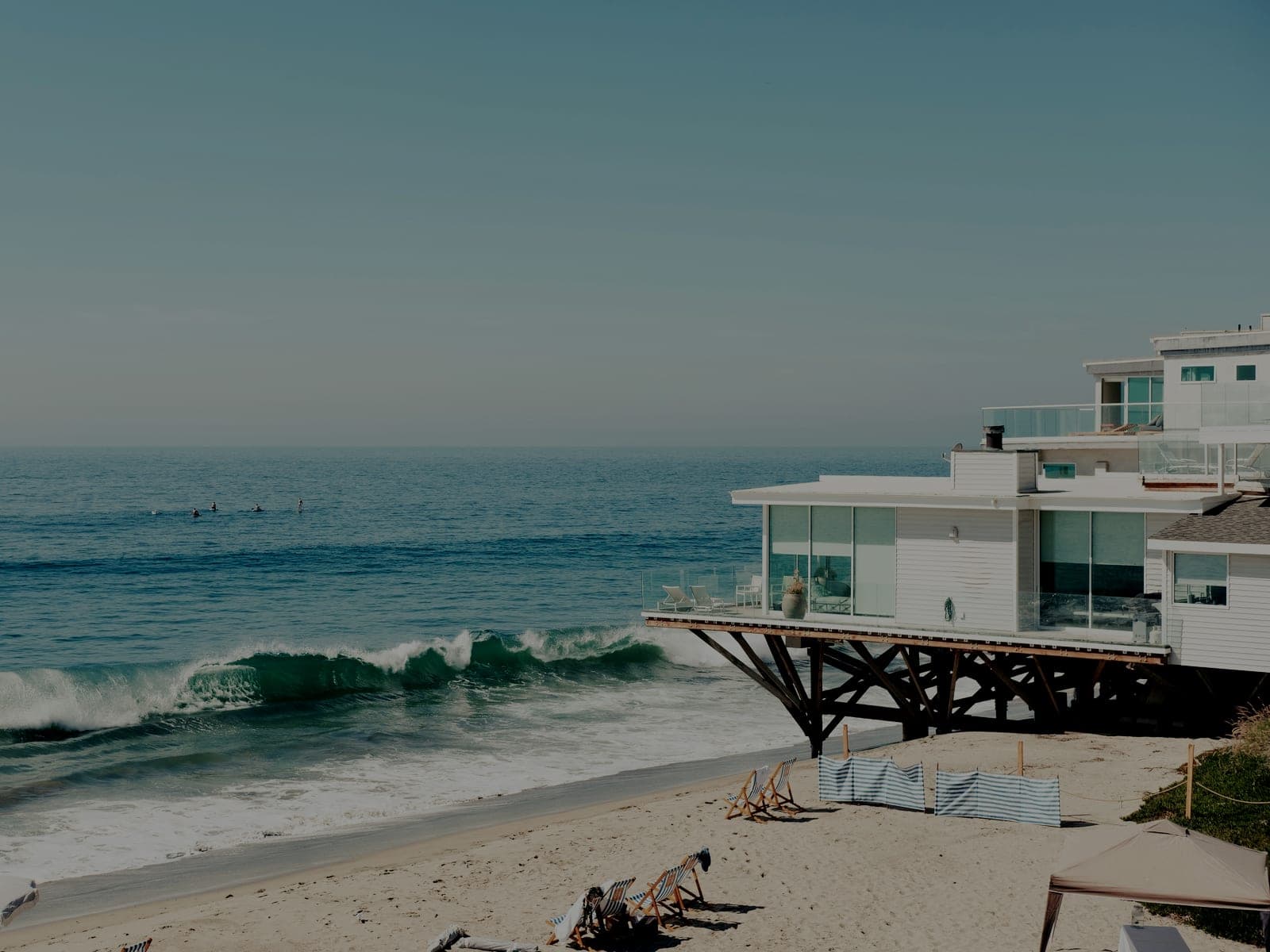 A modern beachfront house built out over the sand on wooden pilings, with surfers in the water beyond it