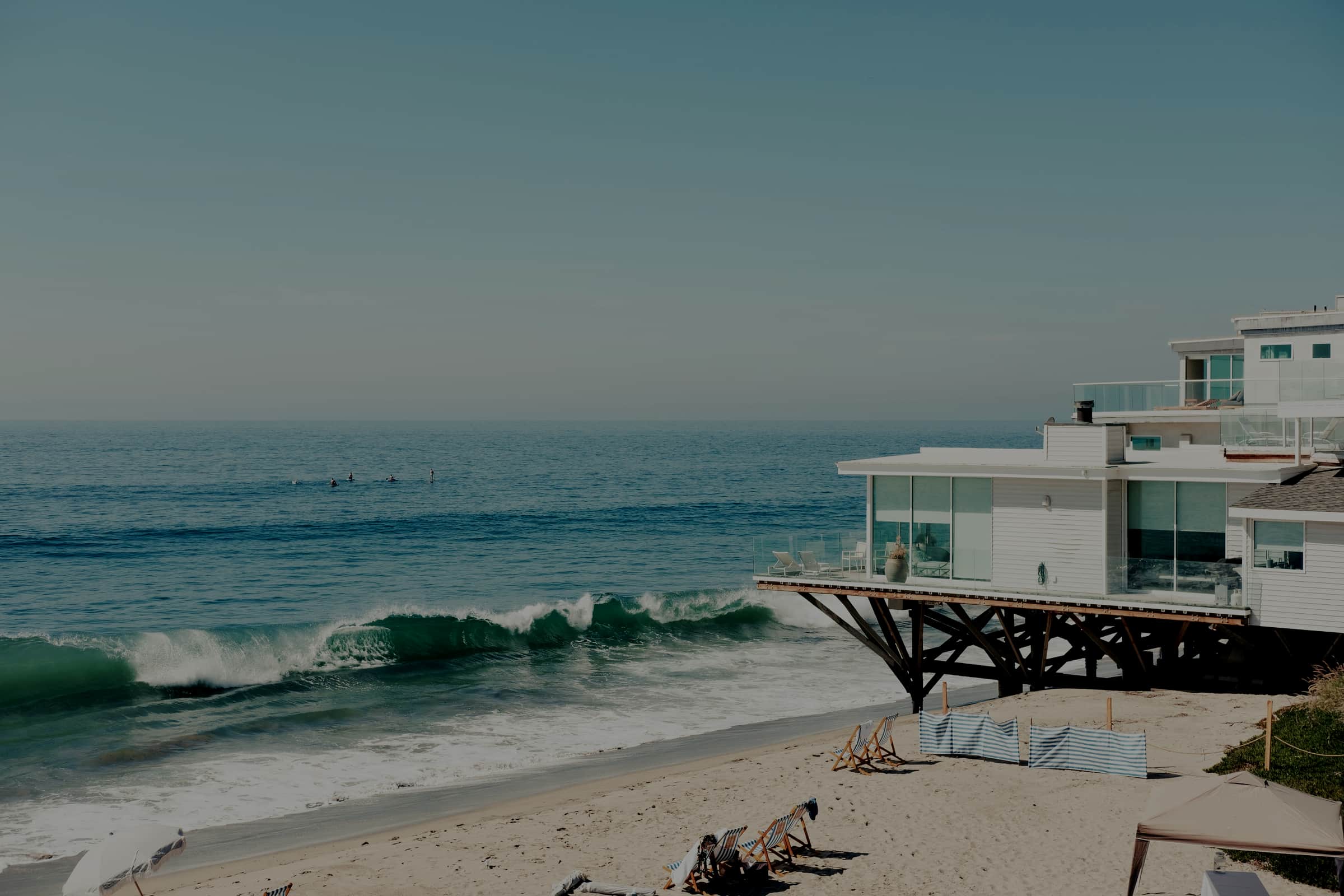 A modern beachfront house built out over the sand on wooden pilings, with surfers in the water beyond it