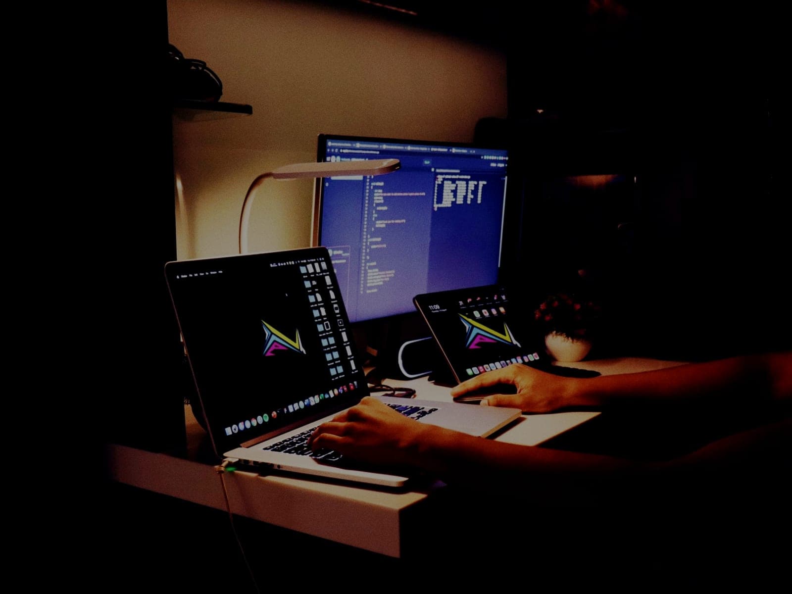 A developer working at night across a laptop, an external monitor showing code, and a tablet