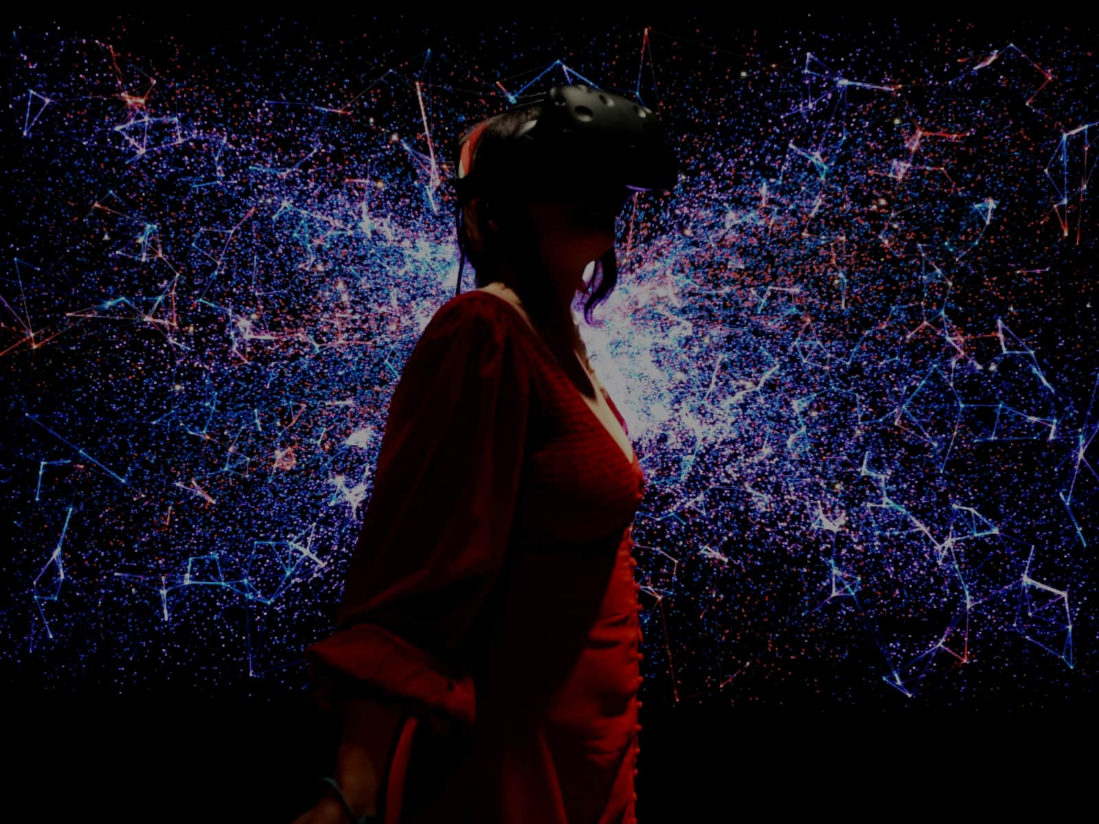 A woman in a red dress wearing a VR headset, standing in front of a wall of blue and pink particle light