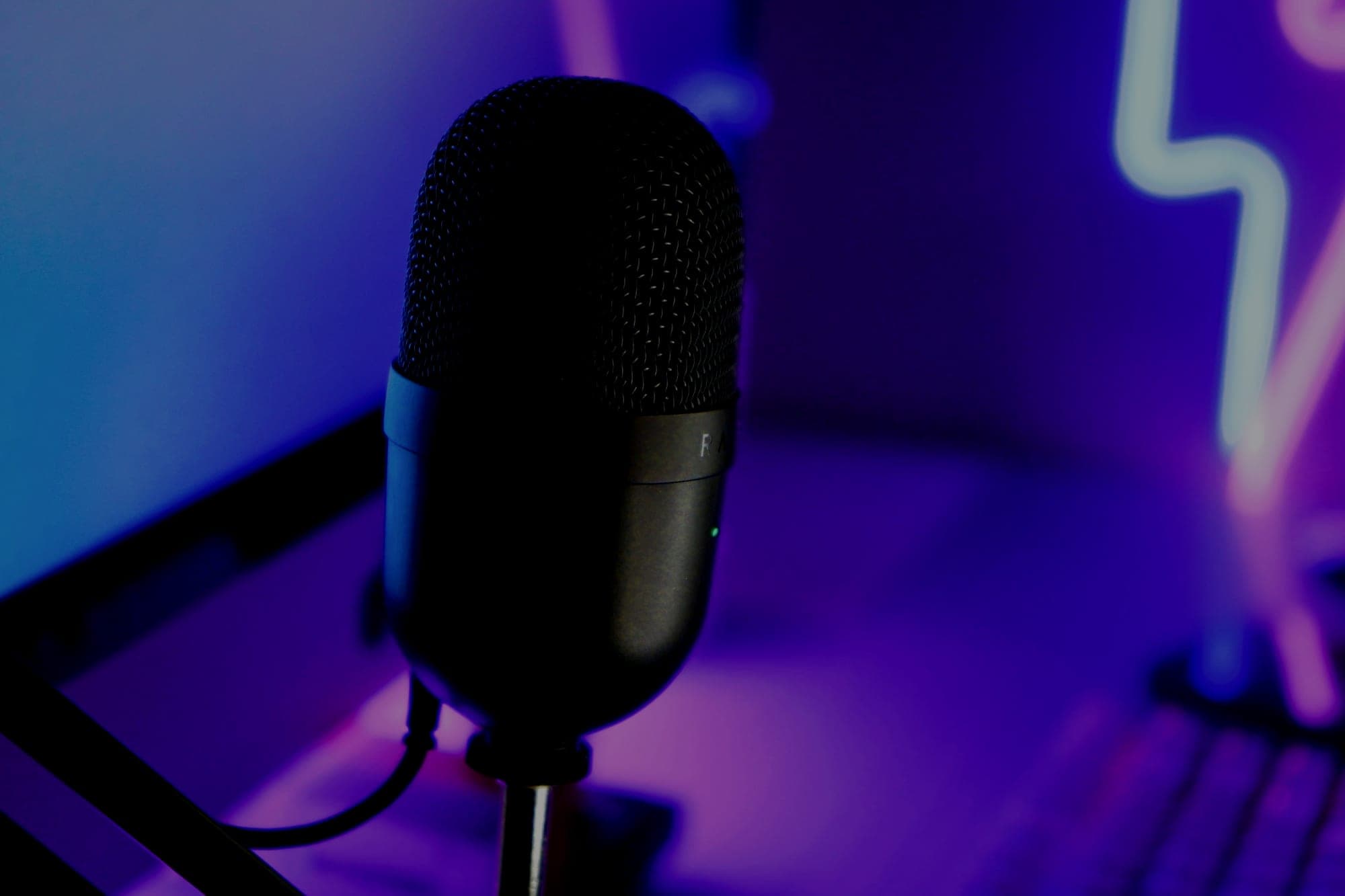 A black condenser microphone on a desk stand, lit purple and blue, with a laptop and neon sign behind it