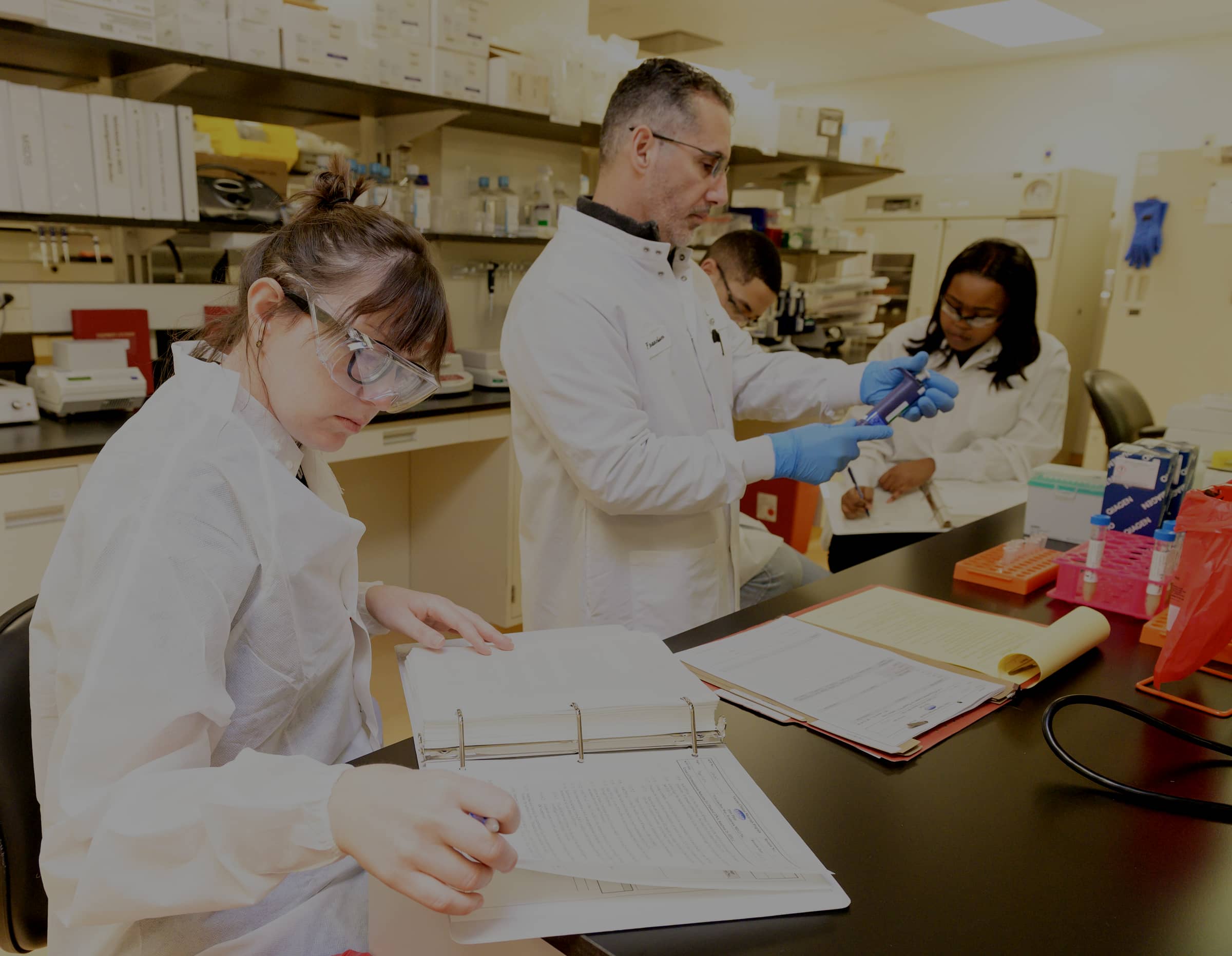 Four researchers in white coats and safety glasses working at a laboratory bench