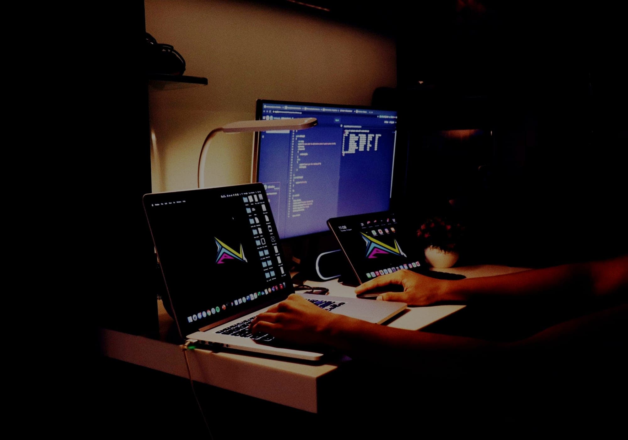 A developer working at night across a laptop, an external monitor showing code, and a tablet