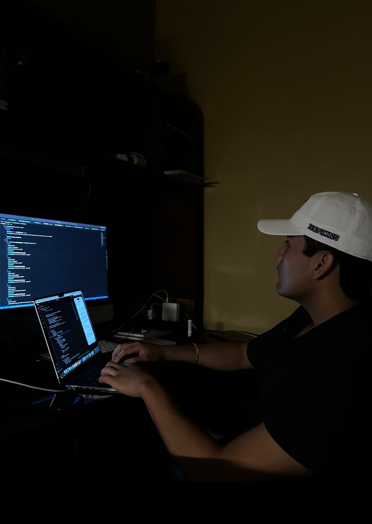 Jeffrey Escobar coding AltaRealm, an AI world-building game, late at night in August 2025, wearing a Johns Hopkins cap